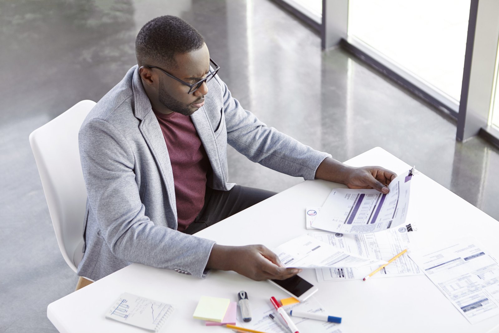 Top view of serious confident young dark skinend male economist, uses paper documents, checks monthly report, has confident expression, sits at his cabinet. People, job and business concept.